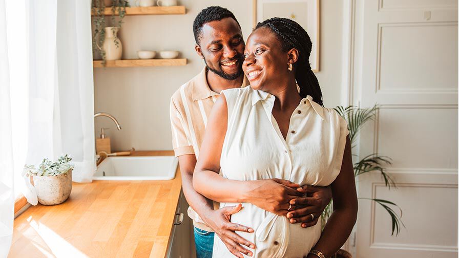Casal feliz em casa a abra&ccedil;ar-se durante a gravidez, mulher gr&aacute;vida segura a barriga com carinho.